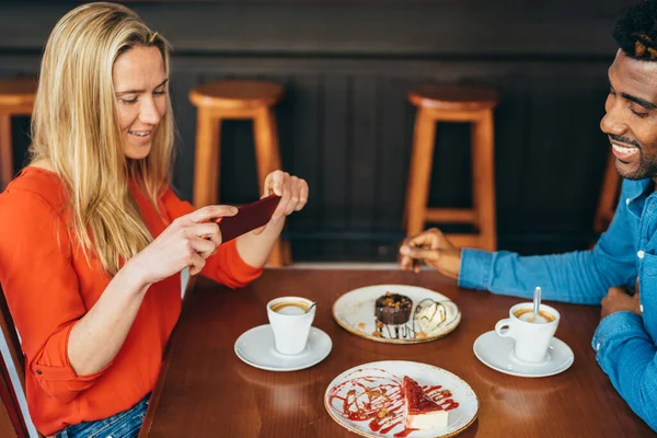 Mujer caucásica tomando una foto con su celular de una tarta de queso mientras bebe café con su novio — Foto de Stock