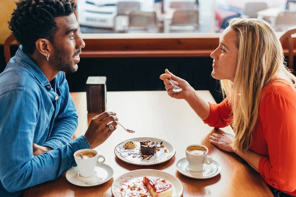 Pareja interracial tomando café y pastel sentado en una mesa en una cafetería — Foto de Stock