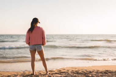 full shot of a woman in sweatshirt and cap watches the sunset on the beach