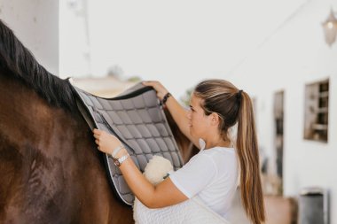 young woman placing a blanket over the back of a horse to fit a saddle. Horseback riding