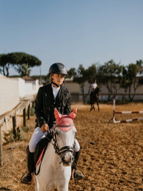 14 year old teenager jogging white horse with pink earmuffs on a riding arena.