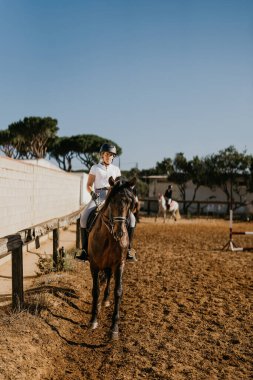 vertical photo of a young woman warming up with her brown horse in the riding arena. Horseback riding school