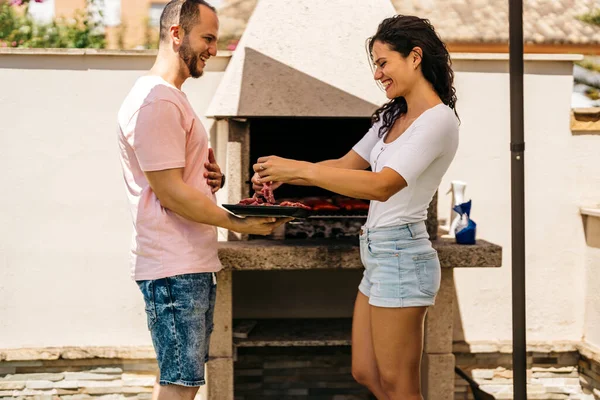 smiling latin woman takes meat from a tray held by a young man to put it on the barbecue. food