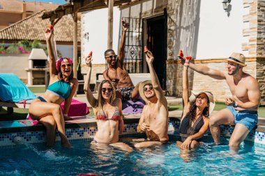 Yüzme havuzunda bir dilim karpuzla kadeh kaldıran bir grup arkadaş. Tatil