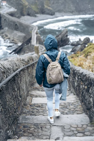 woman walking down the stairs of San Juan de Gaztelugatxe with a raincoat and a backpack