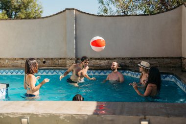 Adam kafası arkadaşlarıyla çevrili bir havuzda plaj topuna vuruyor. Tatil
