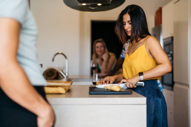 Etrafı arkadaşlarıyla çevrili, ekmek dilimleyen Latin bir kadın.