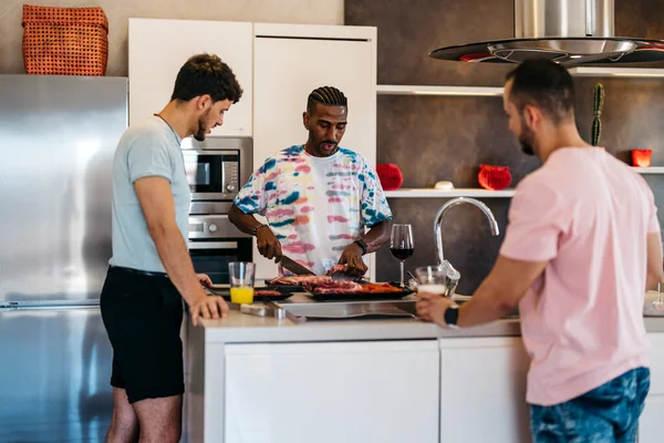 三个朋友在厨房里交谈 一个在用刀切肉 — 图库照片