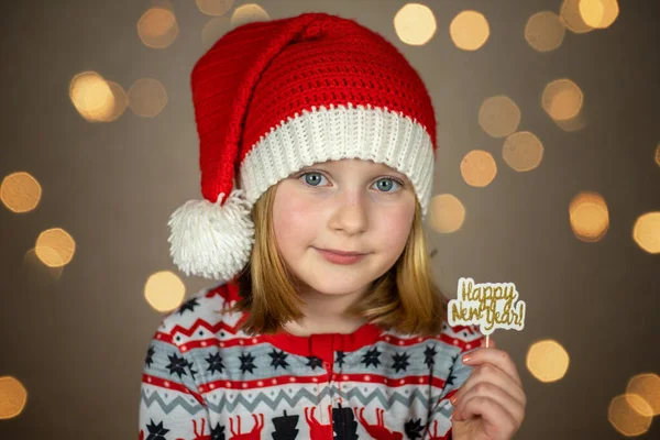 Uma menina em um chapéu de santa detém um sinal de ano novo feliz em um fundo bokeh ano novo — Fotografia de Stock