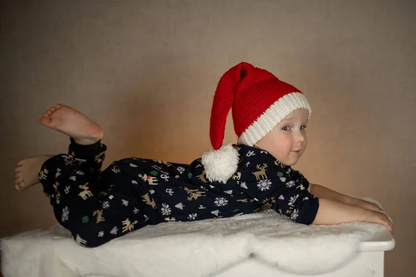 Um pequeno menino bonito em um chapéu de Papai Noel e pijama de Ano Novo encontra-se em um tapete de pele em um fundo liso — Fotografia de Stock