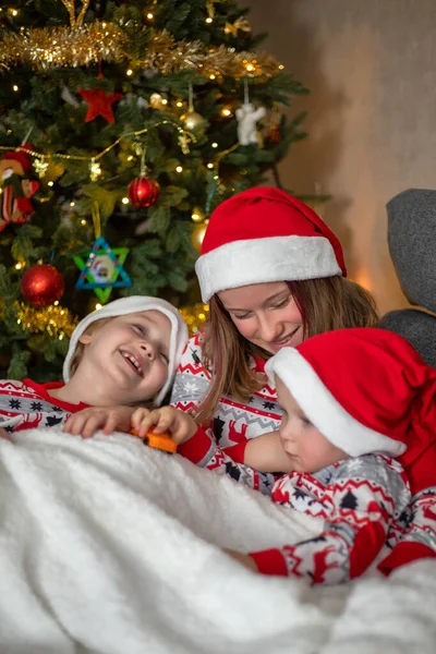 Three children have fun on the background of the Christmas tree. children are dressed in New Years pajamas and santa hats — Stock Photo, Image