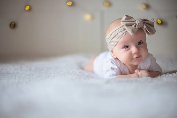 Menina bebê 3 meses de idade encontra-se em uma cama com roupas brancas no estômago e sorri, olha para a câmera, bebê de manhã, conceito de coisas de bebê — Fotografia de Stock