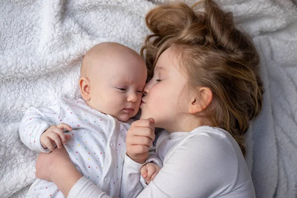Happy children, toddler and older sister, hugging at home on a white blanket, smiling, shot from above. blonde girl hugging her newborn sister.