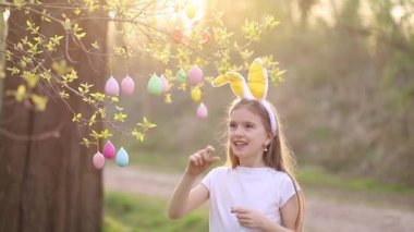 Tavşan kulaklı mutlu bir kız gün batımında bir ağacı paskalya yumurtasıyla süslüyor ve gülüyor. Çok renkli yumurtalar dallara tutunur. Mutlu Paskalyalar