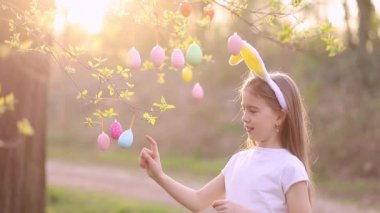 Tavşan kulaklı mutlu bir kız gün batımında bir ağacı paskalya yumurtasıyla süslüyor ve gülüyor. Çok renkli yumurtalar dallara tutunur. Mutlu Paskalyalar