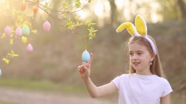 Tavşan kulaklı mutlu bir kız gün batımında bir ağacı paskalya yumurtasıyla süslüyor ve gülüyor. Çok renkli yumurtalar dallara tutunur. Mutlu Paskalyalar
