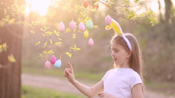 A little happy girl in bunny ears decorates a tree with easter eggs at sunset and laughs. multi-colored eggs hang on branches. happy easter concept — Stock Video