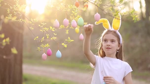 En liten glad flicka i kaninöron dekorerar ett träd med påskägg vid solnedgången och skrattar. flerfärgade ägg hänger på grenar. Glad påsk koncept — Stockvideo