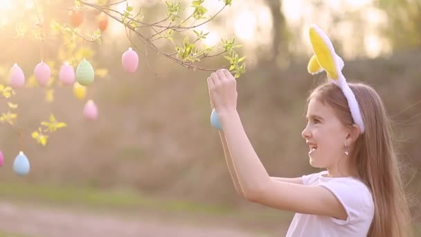 En liten glad flicka i kaninöron dekorerar ett träd med påskägg vid solnedgången och skrattar. flerfärgade ägg hänger på grenar. Glad påsk koncept — Stockvideo