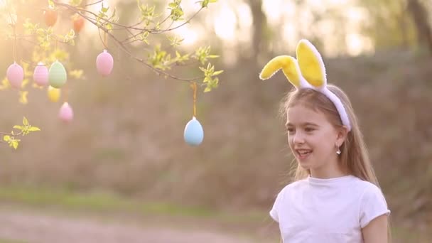 Een klein gelukkig meisje in konijnenoren versiert een boom met paaseieren bij zonsondergang en lacht. meerkleurige eieren hangen aan takken. Gelukkig Pasen concept — Stockvideo