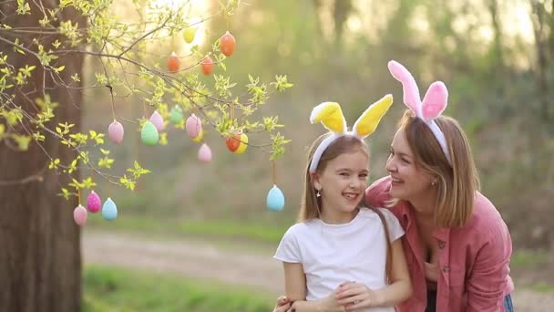 Gelukkige moeder en dochter in konijnenoren vieren Pasen, knuffelen, kussen en lachen op de achtergrond van de zonsondergang. familietraditie van het versieren van een boom met paaseieren voor Pasen — Stockvideo