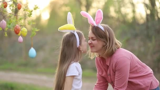 Feliz mamá e hija en orejas de conejo celebrando la Pascua, abrazándose, besándose y frotándose la nariz contra el fondo de la puesta de sol. tradición familiar de decorar un árbol con huevos de Pascua — Vídeos de Stock