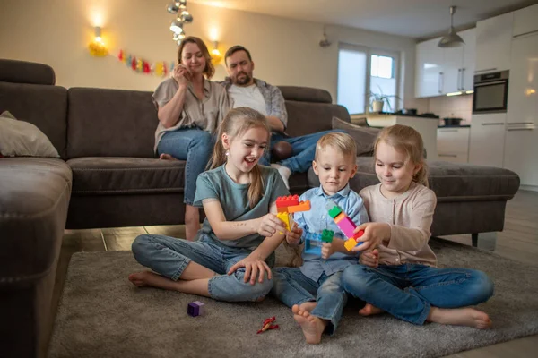 Kinderen broer en zussen veel plezier spelen op de vloer thuis terwijl ouders ontspannen op de bank — Stockfoto