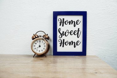 Home Sweet Home tipografisi ahşap masa ve beyaz duvar arkaplanı üzerindeki alarm saati