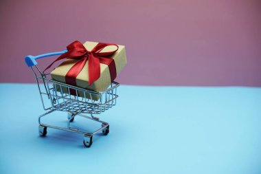 Alışveriş arabasında pembe ve mavi arka planda kırmızı kurdeleli hediye kutusu.