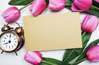 Boş kağıt kart, pembe lale çiçeği ve çalar saati olan tahta arka planda yatıyordu.
