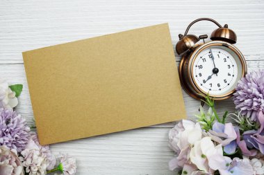 Çalar saati ve çiçek buketi düzlüğü olan boş kağıt kart tahta arka planda yatıyordu.