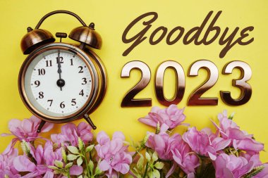 Goodbye 2023 text with alarmclock and flower decoration on yellow background