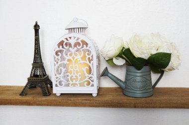 Ev dekorasyonu aksesuarları Eyfel heykeli, LED mum ışığında beyaz fener ve ahşap raflarda metal vazoda çiçek ve beyaz beton duvar.