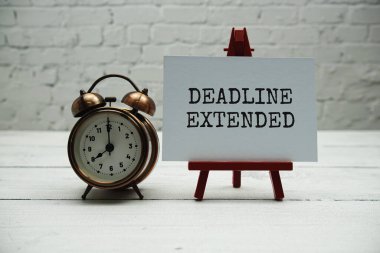 Deadline Extended text and alarm clock on white brick wall and wooden background