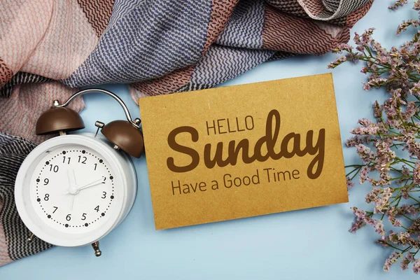 Hello Sunday Have a good Time written on paper card with flower and alarm clock on blue background