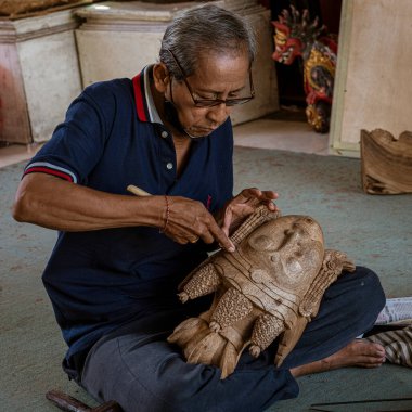 Man sits cross-legged on floor while finishing carving wooden mask.