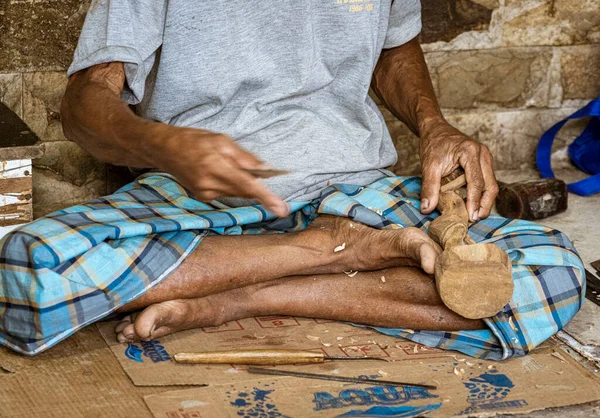 Man sits cross-legged while carving wooden statue.