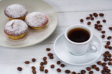 Cupcakes sprinkled with powdered sugar on a plate and a cup of coffee with coffee beans on a white wooden table