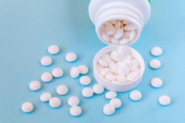 Mavi arka planda kapağı olan beyaz yuvarlak haplar şişeden yayılmış.