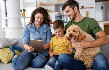Modern aletleri olan mutlu genç bir aile ve evde oynayıp eğlenen bir köpek.