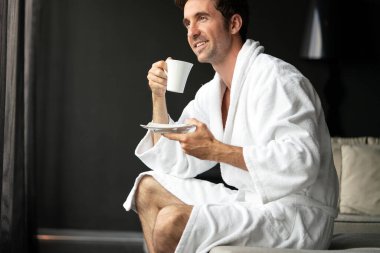 Genç, mutlu yakışıklı adam sabahları bornozlu kahve içiyor.