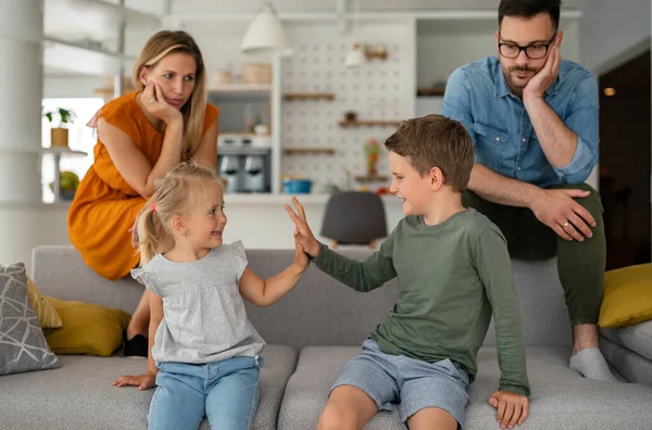 Tired parents sitting on couch feels annoyed exhausted while happy children playing together at home