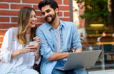Kafede oturmuş gülümseyen, dizüstü bilgisayara bakan, keyifle vakit geçiren, kahve içen mutlu genç bir çift..