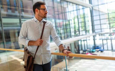 Cam binanın önünde duran, resmi kıyafetler giymiş, gülümseyen genç ve yakışıklı bir iş adamının portresi.