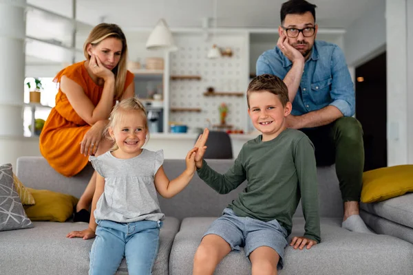 Tired parents sitting on couch feels annoyed exhausted while happy children playing together at home