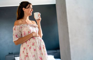 Güzel, mutlu, genç, hamile bir kadın evde gülümseyip kahve, çay ya da vitamin içiyor.