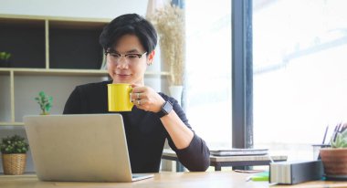 Genç Asyalı adam kahve içiyor ve dizüstü bilgisayarda çalışıyor..