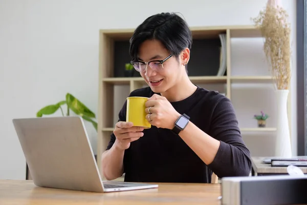 笑着拿着咖啡杯的亚洲年轻人 在笔记本电脑上阅读新闻 — 图库照片