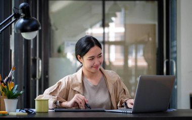 Gülümseyen kadın yaratıcı tasarımcı modern iş yerinde oturuyor ve dizüstü bilgisayarla çalışıyor..