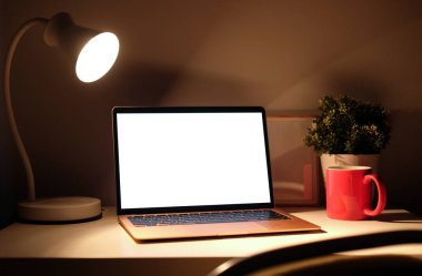 Bilgisayar laptopunu boş ekran ve parlayan lambayla karanlık odadaki ahşap masaya yapıştır.